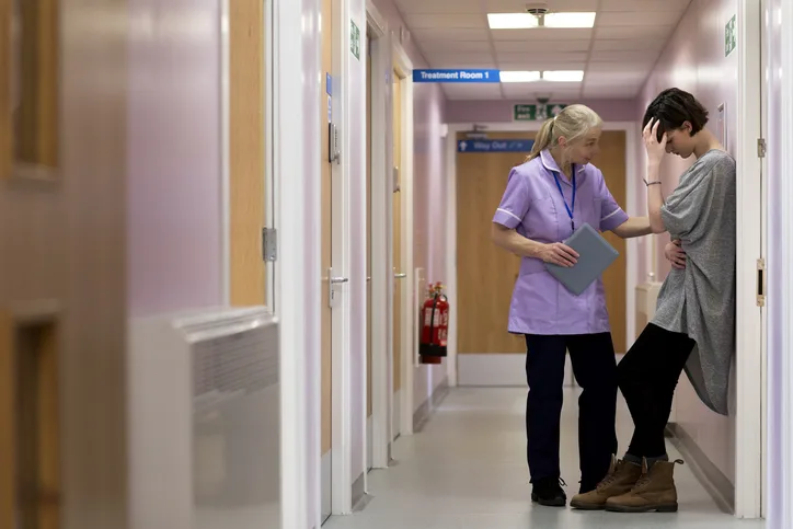 Medical woman comforts young female adult as she stands with her back against to the wall in a hospital corridor